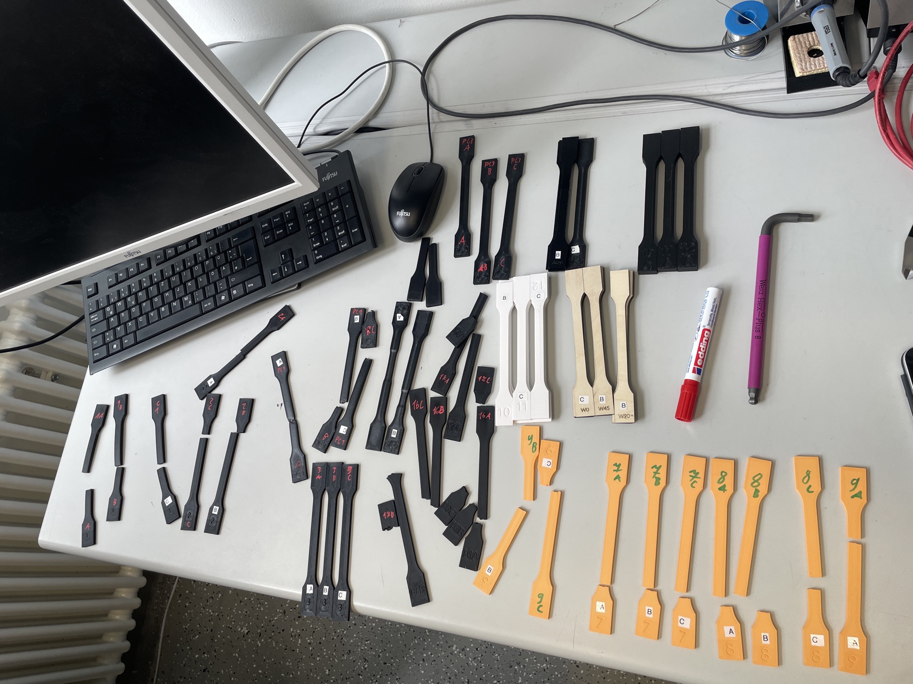 Dozens of labelled 3D-printed tensile test specimens on a lab bench, many broken at the neck